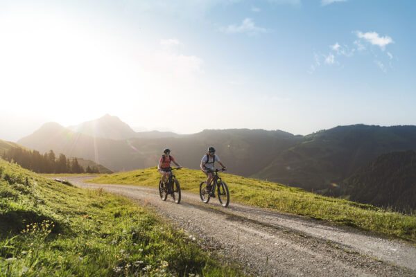 Zwei Radfahrer fahren auf einem Schotterweg durch eine sonnige Berglandschaft des Pillerseetals.