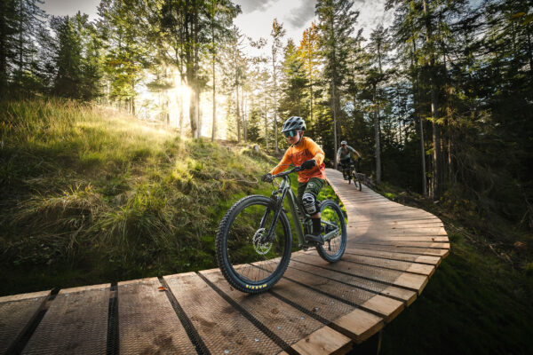 Zwei Mountainbiker, darunter ein Kind, fahren über einen Holzsteg im Wald bei Sonnenlicht.
