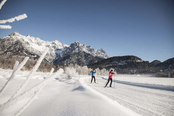 Zwei Langläufer gleiten bei Sonnenschein durch eine tief verschneite Winterlandschaft mit imposantem Bergpanorama.