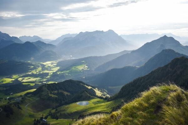Weitläufiger Blick über eine grüne Berglandschaft mit sanften Hügeln und beeindruckenden Gipfeln des Wilden Kaisers in der Ferne.
