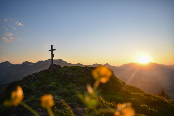 Sonnenaufgang am Gipfel mit Gipfelkreuz und Blick über eine beeindruckende Berglandschaft.