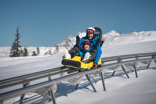 Vater und Kind fahren lachend mit einem Coaster durch eine verschneite Winterlandschaft.