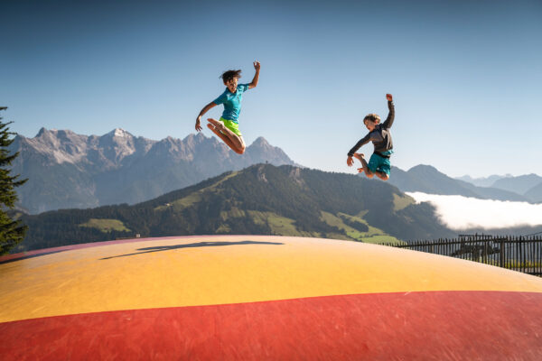 Zwei Kinder springen ausgelassen auf einem großen Luftkissen vor einer beeindruckenden Bergkulisse.