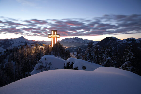 Beleuchtetes Jakobskreuz auf der Buchensteinwand im tief verschneiten Winterpanorama bei Sonnenuntergang mit Blick auf die Kitzbüheler Alpen.