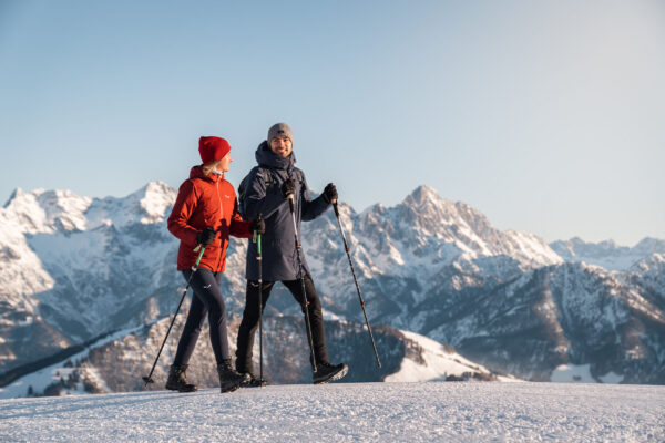 Zwei Winterwanderer mit Schneeschuhen genießen bei Sonnenschein den Blick auf die verschneiten Gipfel der Alpen im Pillerseetal.