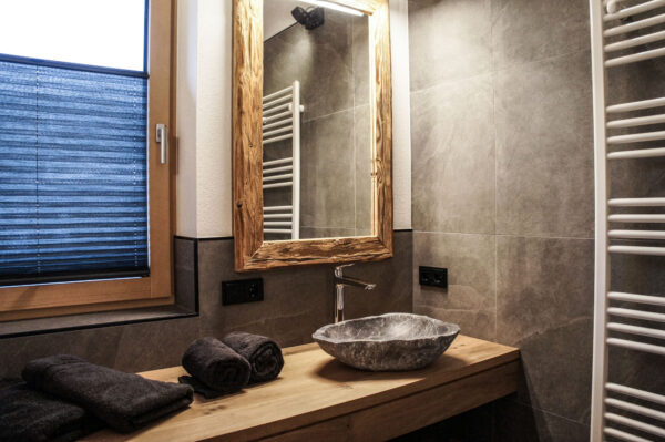 Modernes Badezimmer mit Naturstein-Waschbecken auf rustikaler Holzplatte, großem Holzrahmen-Spiegel und seitlichem Fenster mit Plissee. Rechts im Bild ein weißer Handtuchheizkörper, auf der Ablage liegen gerollte Handtücher.