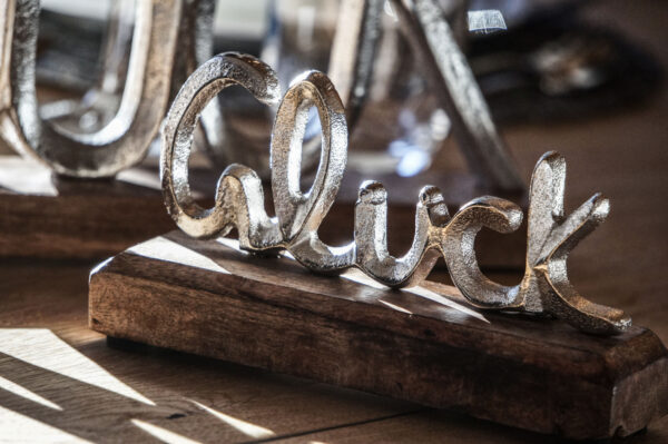 Nahaufnahme einer silbernen Dekoskulptur mit dem Schriftzug „Glück“ auf einem Holzsockel, stilvoll arrangiert auf einem gedeckten Holztisch im Chalet.