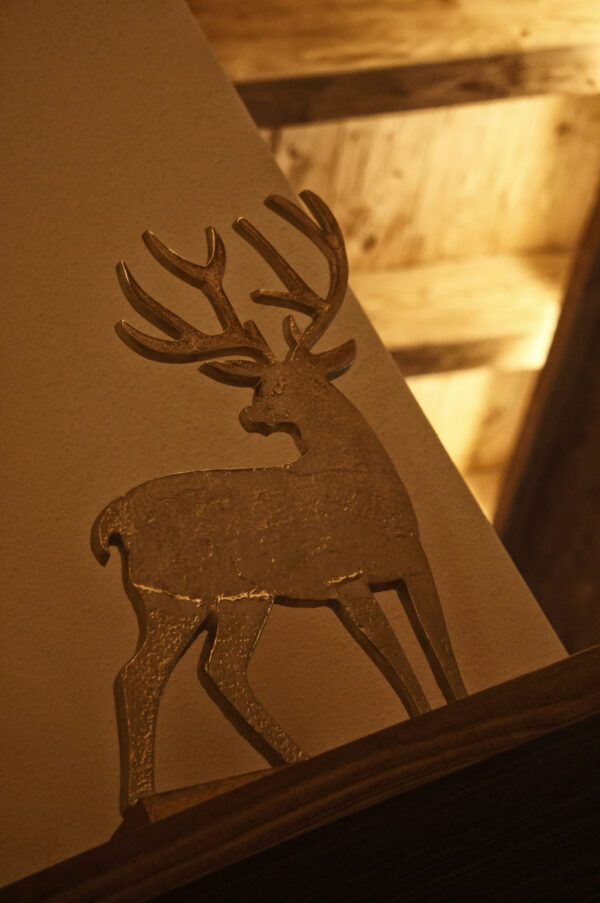 Dekorative Metallfigur eines Hirsches mit Geweih auf einer Wandvorsprung, beleuchtet von warmem Chalet-Licht.