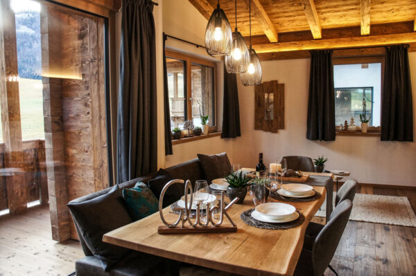 Gemütlicher Essbereich im Chalet-Stil mit großem Holztisch, gedeckt mit stilvollen weißen Tellern, Gläsern und Dekorationen wie Sukkulenten, Kerzen und der Metallschrift „Glück“. Der Raum ist in warmen Tönen gehalten, mit Holzbalkendecke, rustikalem Boden und großen Fenstern mit Blick auf die Natur. Drei moderne Pendelleuchten hängen über dem Tisch, dunkle Vorhänge rahmen die Fenster. Im Hintergrund: eine einladende Sitzbank und Fensterbänke mit Pflanzen.