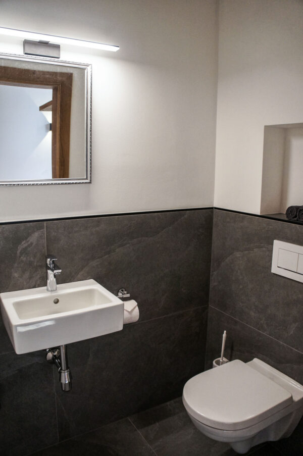 Modernes Gäste-WC mit halbhoch gefliester Wand in dunklem Naturstein-Look, kombiniert mit einer weißen Wand im oberen Bereich. Ein rechteckiges, wandmontiertes Waschbecken mit verchromter Armatur befindet sich unter einem schlichten Spiegel mit dezentem Rahmen. Daneben ist eine wandhängende Toilette platziert. Auf einem seitlichen Wandvorsprung liegen gerollte, dunkle Handtücher.