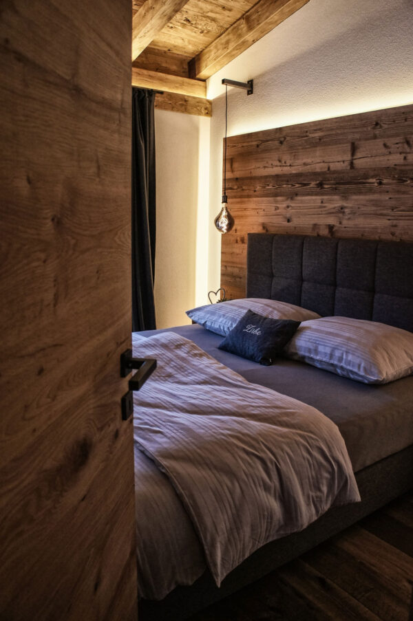 Gemütliches Schlafzimmer mit offenem Holzbalken-Plafond, indirekter Beleuchtung und rustikaler Holzverkleidung hinter dem Kopfteil des Bettes. Modernes Boxspringbett mit grauer Bettwäsche, einem Dekokissen mit der Aufschrift „Zirbe“ und einer stilvollen Hängelampe. Die Tür ist leicht geöffnet und gibt den Blick in den behaglichen Raum frei.
