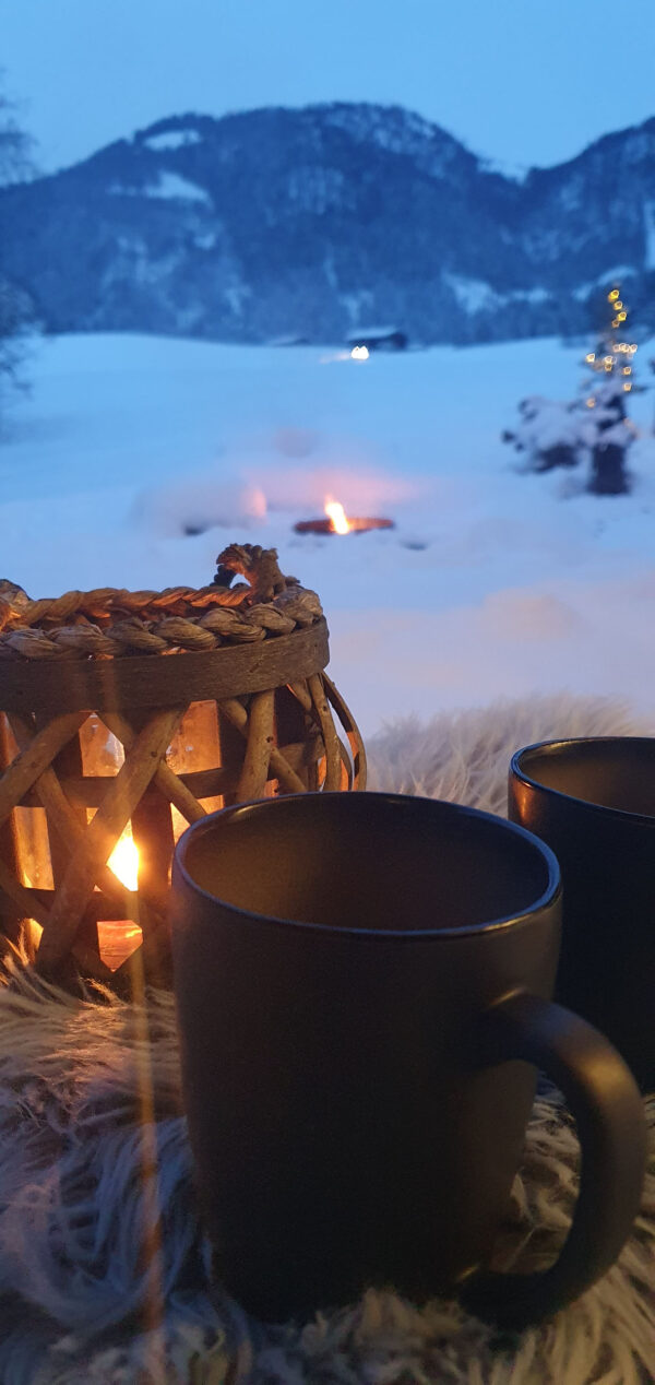 Winterliches Panorama vom Chalet-Apartment Fieberbrunn: auf einer fellbedeckten Terrasse stehen zwei schwarze Tassen neben einer rustikalen Kerzen-Laterne, dahinter lodert ein Lagerfeuer im verschneiten Garten vor der Bergkulisse der Kitzbüheler Alpen – perfekte, romantische Winterurlaub-Atmosphäre in Tirol.