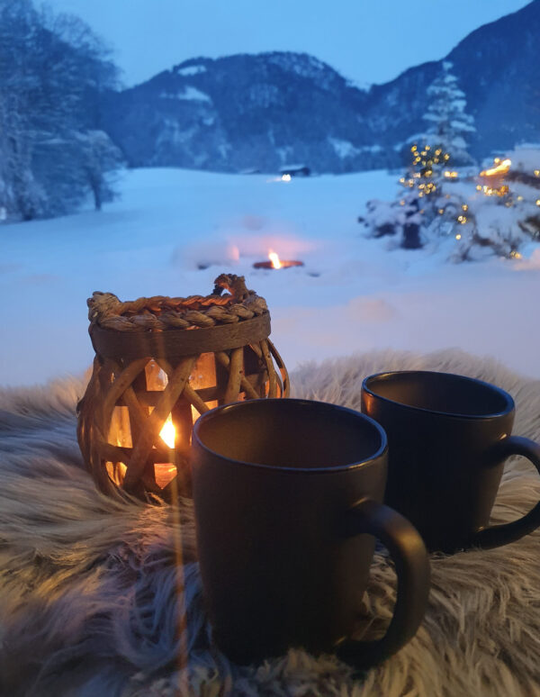 Zwei Teetassen am Lagerfeuer vor verschneitem Bergpanorama