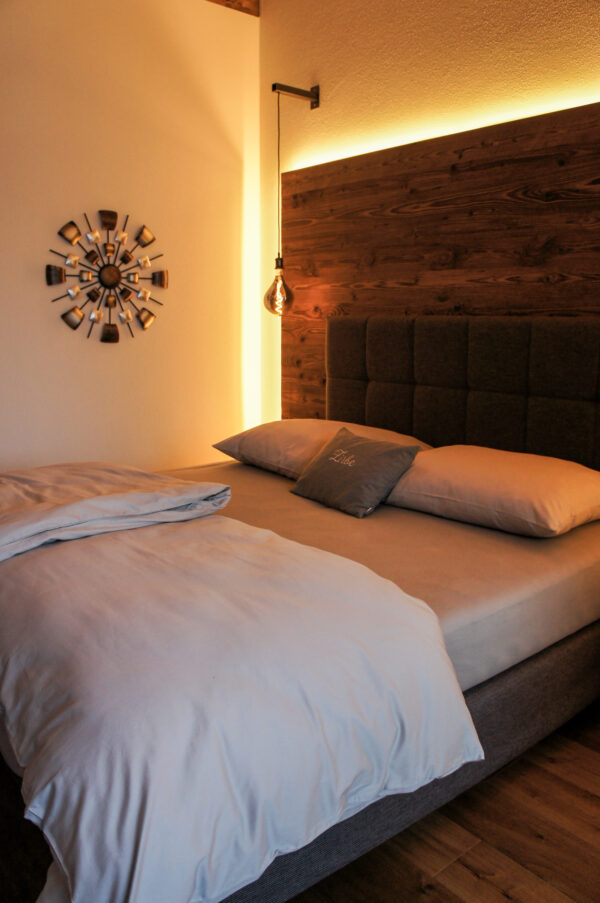 Doppelbett mit Holzrückwand, seitlicher Hängelampe und stilisierter Wanddeko in stimmungsvoll beleuchtetem Schlafzimmer