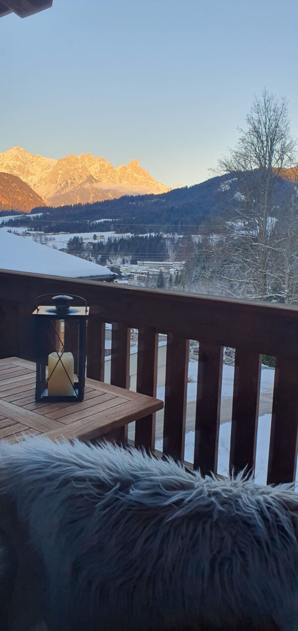 Spätwinterlicher Balkon mit Kerzenlaterne und schneebedeckter Bergkulisse im Abendlicht