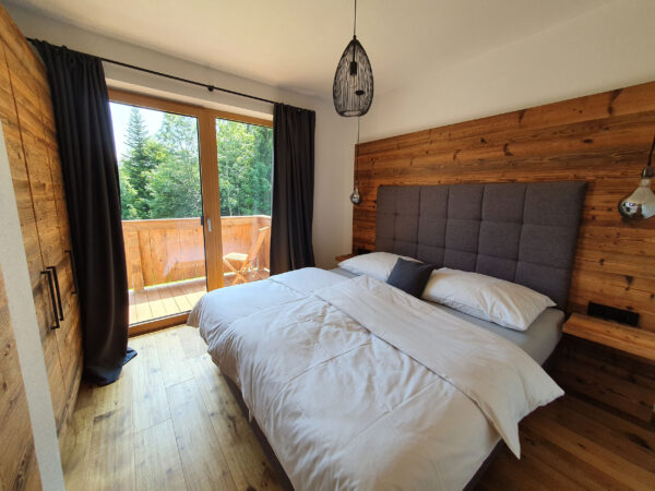 chlafzimmer mit Boxspringbett, grauem gepolstertem Kopfteil, Holzwandverkleidung und Blick auf den Balkon mit Tageslicht.