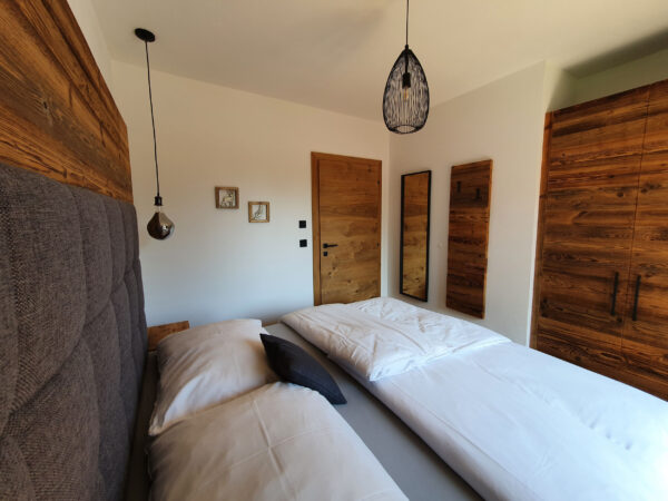 Blick vom Kopfteil des Boxspringbetts in Richtung Tür, zwei dekorative Holzrahmen an der Wand, Spiegel und Hakenleiste aus Holz, rustikaler Kleiderschrank mit schwarzen Griffen.