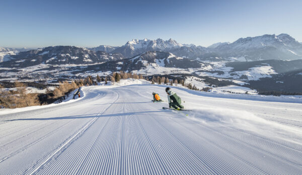 Zwei Skifahrer fahren auf einer breiten, frisch präparierten Piste mit Panoramablick auf die umliegenden Berge.